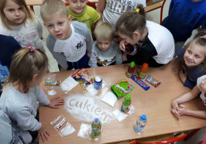 Dzieci siedzą przy stoliku, na którym są woreczki z cukrem, jaki znajduje się w niektórych produktach